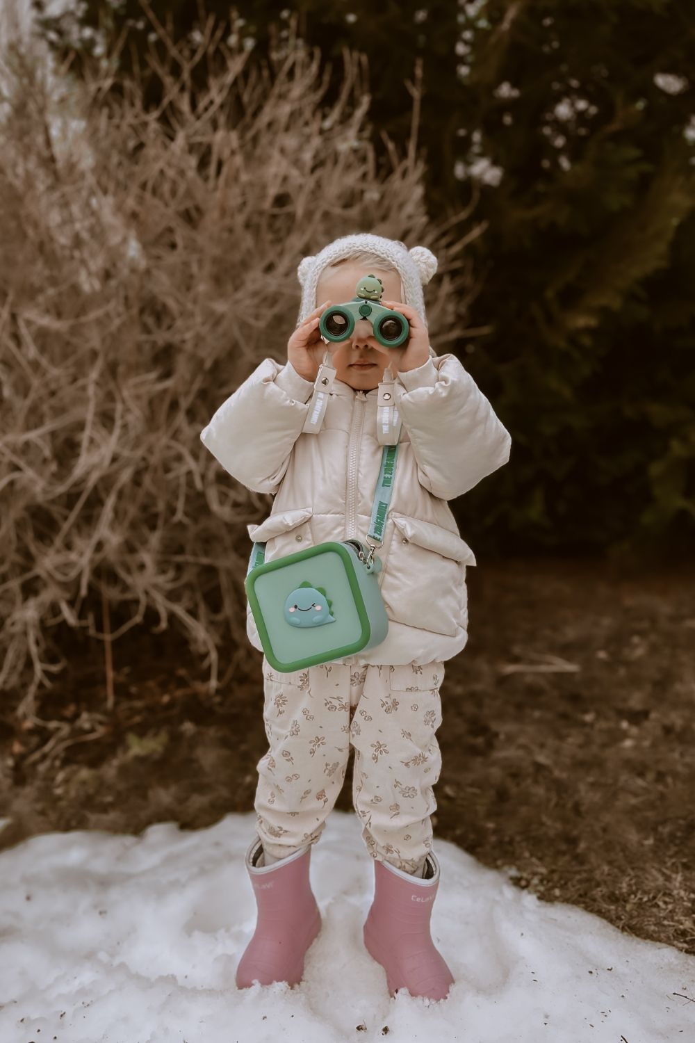 dino green silicon bag, child is carrying the bag around its neck, while playing with a pair of binoculars, standing in the snow