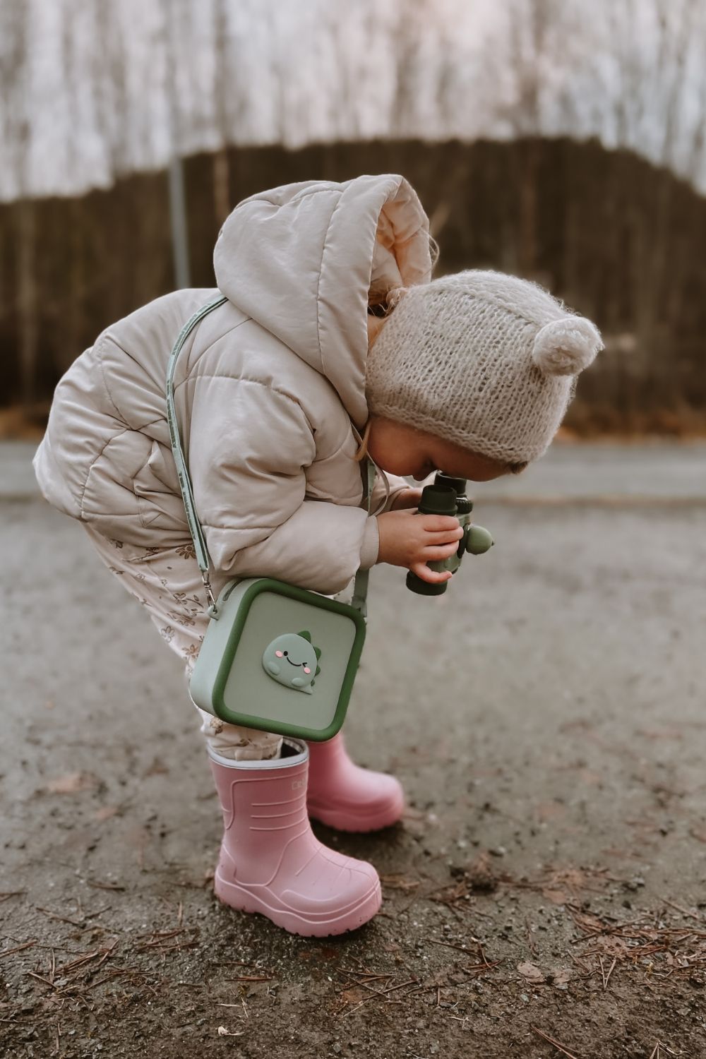 dino green silicon bag, child is playing outside with a pair of binoculars, ooking at wood and stones