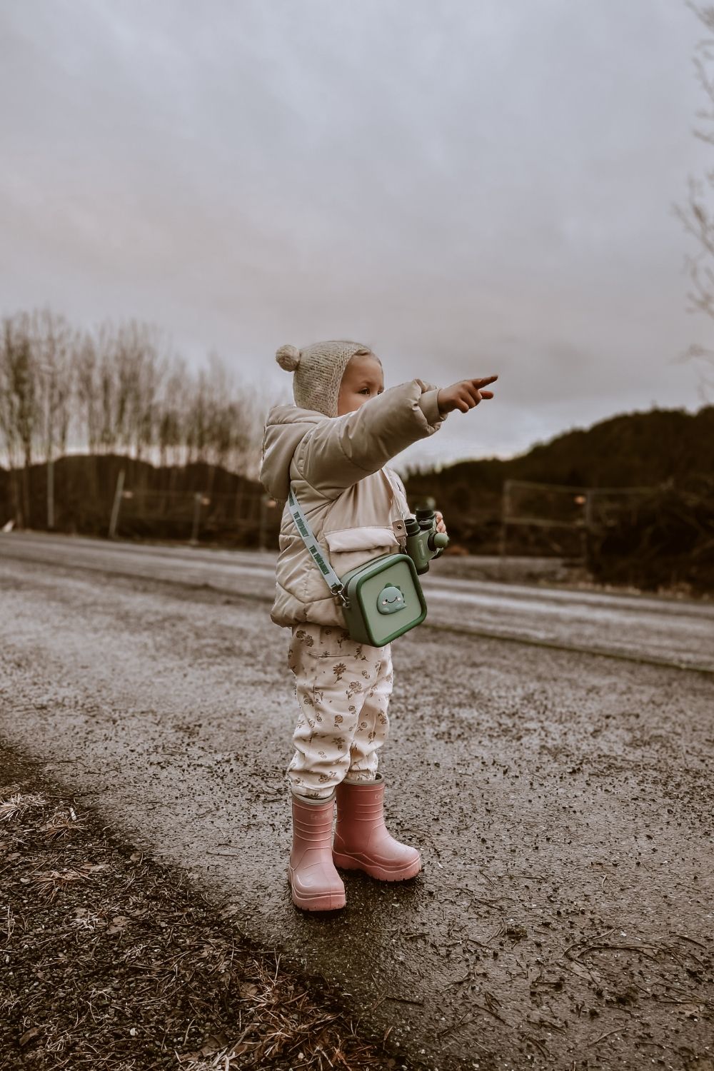 dino green silicon bag, child is carrying the bag around its neck, standing outside, pointing at a place, having a pair of binoculars in its hand