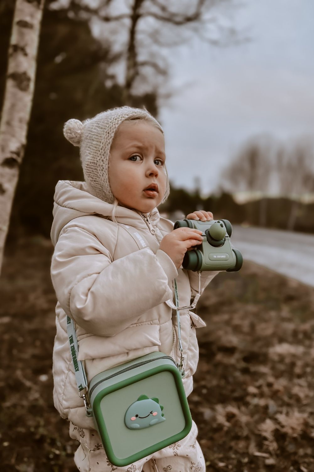 dino green silicon bag, child is carrying the bag around its neck, holding a pair of binoculars, standing outside next to a tree