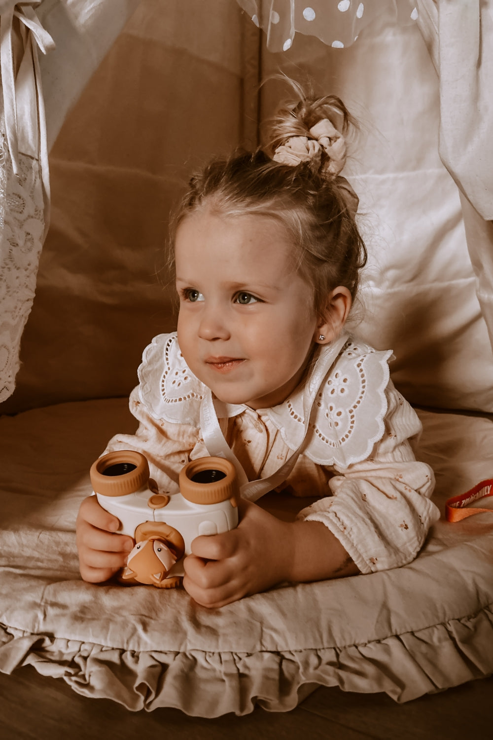 The girl is smiling and lying on the playmat, holding the binoculars at the ready.
