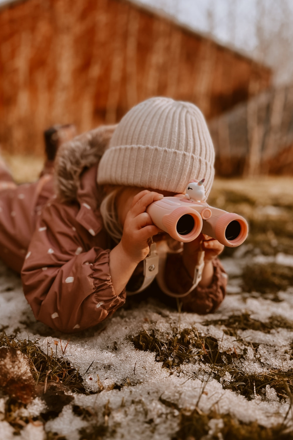 The girl is lying on the barely snow-covered grass, propped up on her elbows, looking through binoculars.