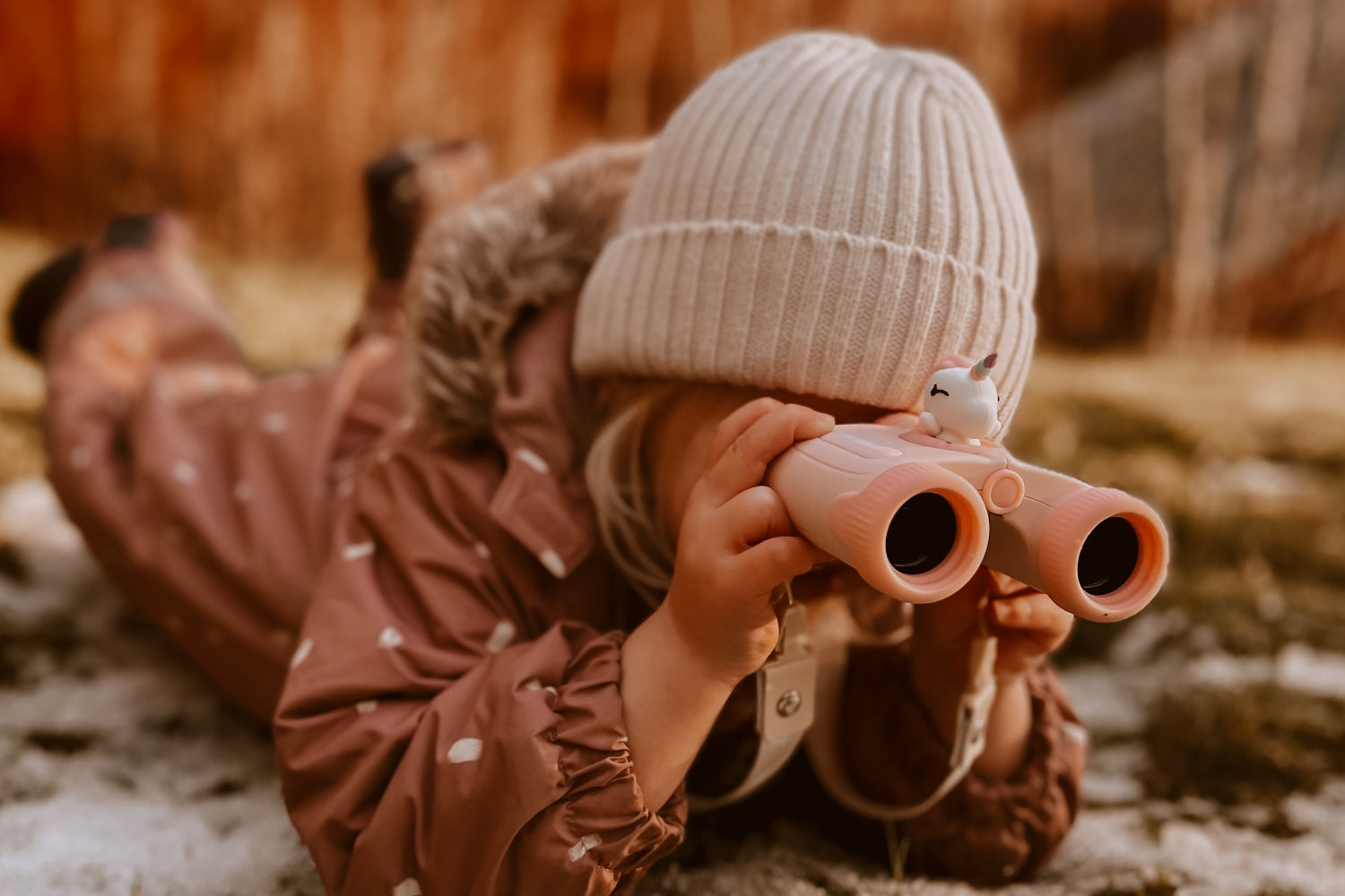 The girl is lying on the barely snow-covered grass, propped up on her elbows, looking through binoculars.