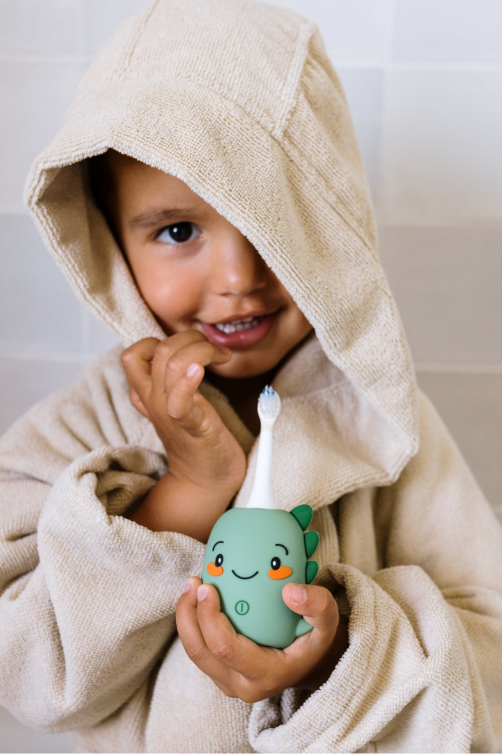 A smiling child wrapped in a beige hooded towel holds dino green toothbrush