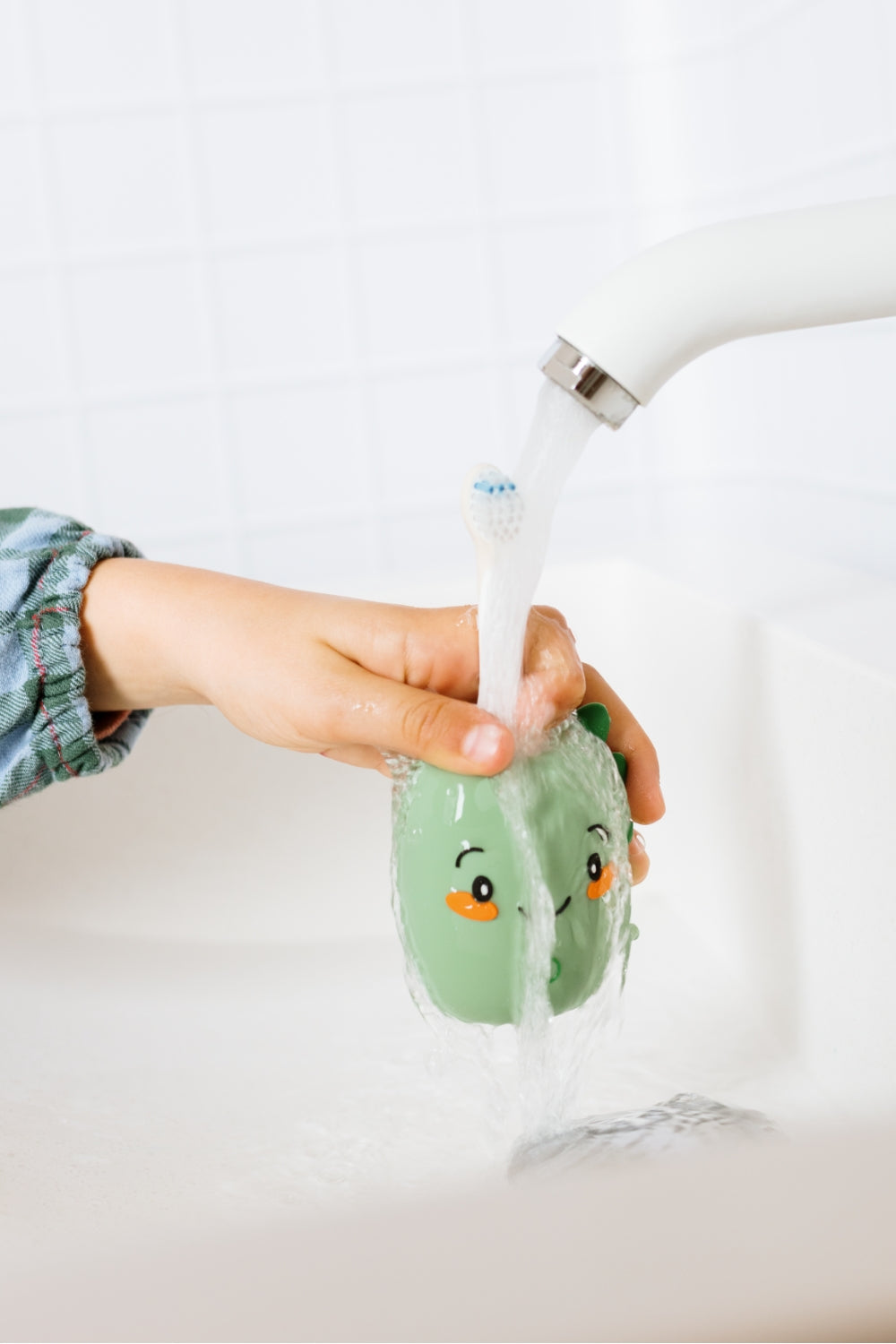 A dino toothbrush  with running water from a faucet in a white sink. A child's hand are visible holding the product.