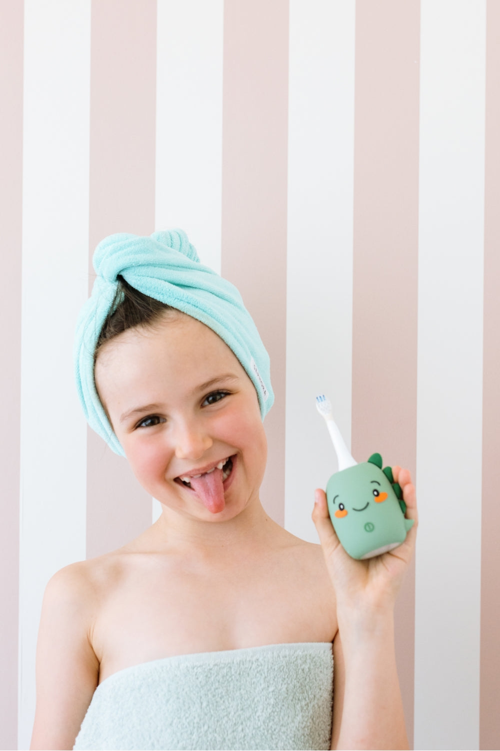 A smiling child with a mint green towel wrapped around their head and a matching towel strap across their shoulders holds a small dino toothbrush.