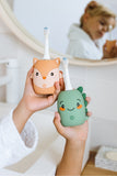 odalTwo cute cartoon electric toothbrushes with colorful silicone covers are held in a person's hands in a bright bathroom setting. One toothbrush has a fox , and the other has a green dinosaur.