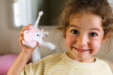 odalA smiling young girl with curly brown hair holds a pink unicorn-shaped electric toothbrush unicorn in her hand