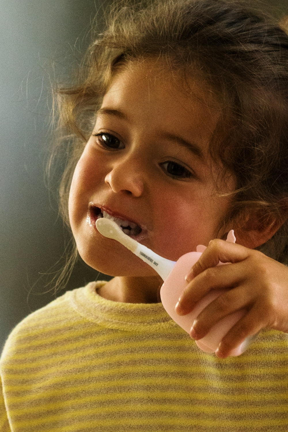 A young child with curly brown hair brushing their teeth with a unicorn toothbrush.