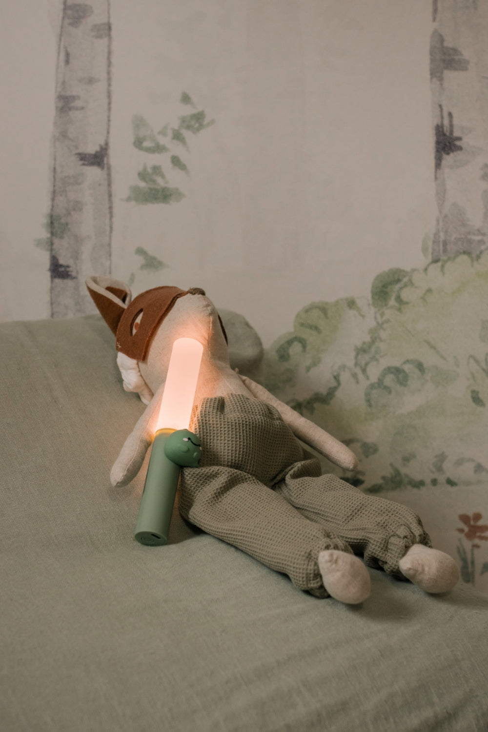 A stuffed animal with a lamp on a bed in a room with a forest-themed wall.